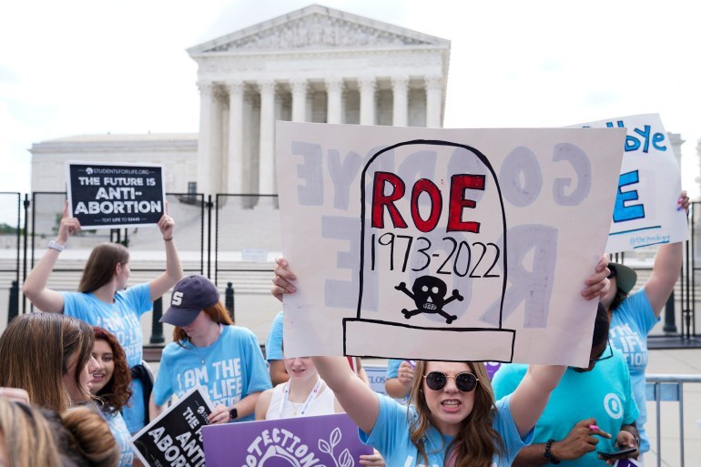 A woman holds up a hand-drawn poster that depicts a tombstone with the words "Roe 1973-2022" written on it. The Supreme Court is behind her.