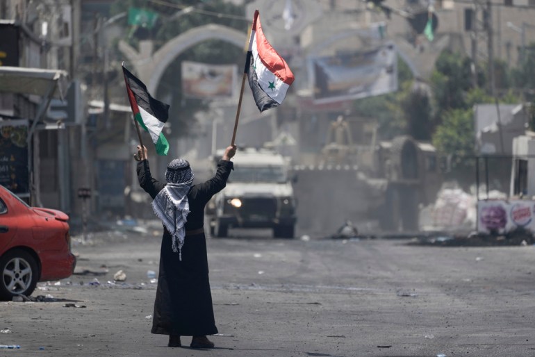 A Palestinian man waves Palestinian and Syrian flags in front of an Israeli army vehicle during a military raid in the Jenin refugee camp, a militant stronghold, in the occupied West Bank, Tuesday, July 4, 2023. Palestinian health officials said at least 10 Palestinians were killed in the operation, which began Monday. (AP Photo/Majdi Mohammed)