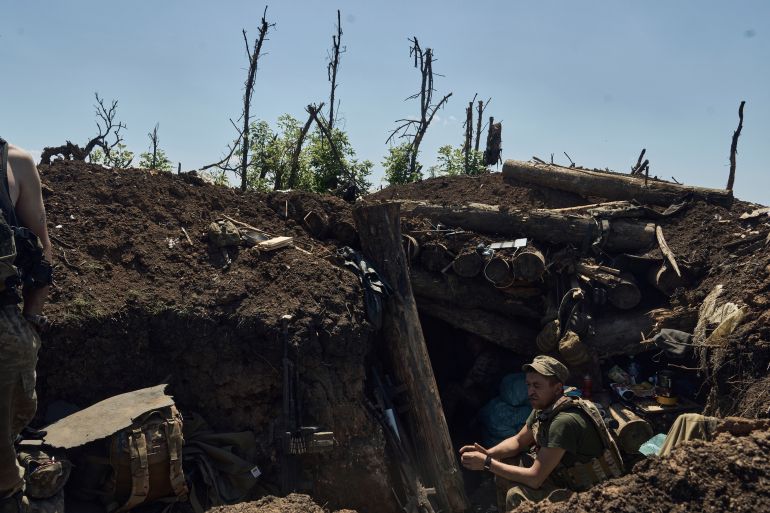 A Ukrainian soldier sitting in a recently taken Russian trench near Bakhmut. There is lots of dark mud and broken trees around.