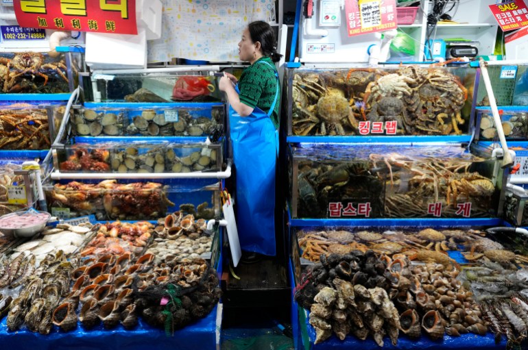 A woman standing between stalls laden with seafood in a Seoul market. She is wearing blue overalls.