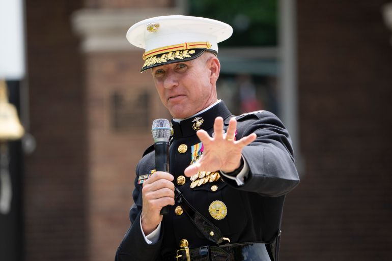 A man raises a hand to gesture as he speaks into a mic. He wears a marine dress uniform.