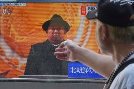 A TV screen showing a pictire of Kim Jong Un in a black trilby and dark coat. Someone is pointing at the screen.