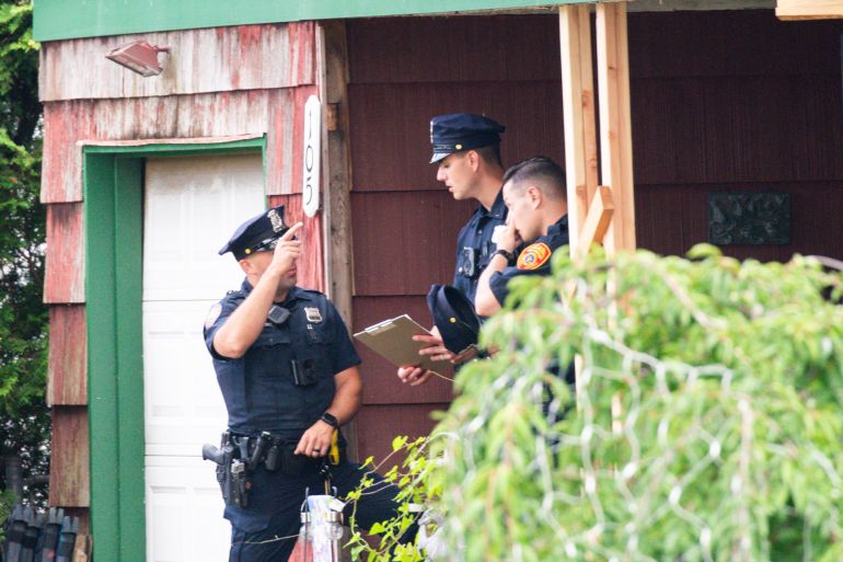 A police officer on a porch points while another two officers listen to him speak. The house is red with wood panels and green trim. A white garage door is visible.