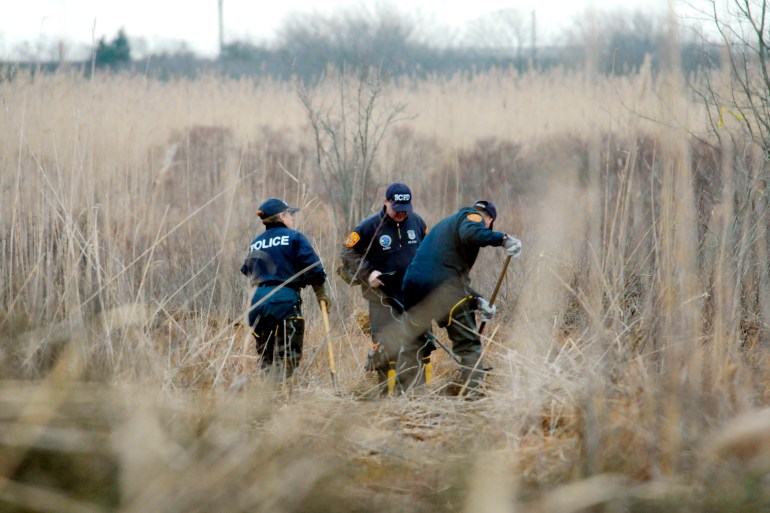 Amidst the tall dry reeds of a swamp, three police officers in black with a metal detector on the ground.