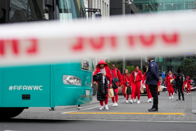 Sebuah tim sepak bola, berpakaian merah, berkendara menuju bus berwarna aqua yang dikelilingi pita peringatan.