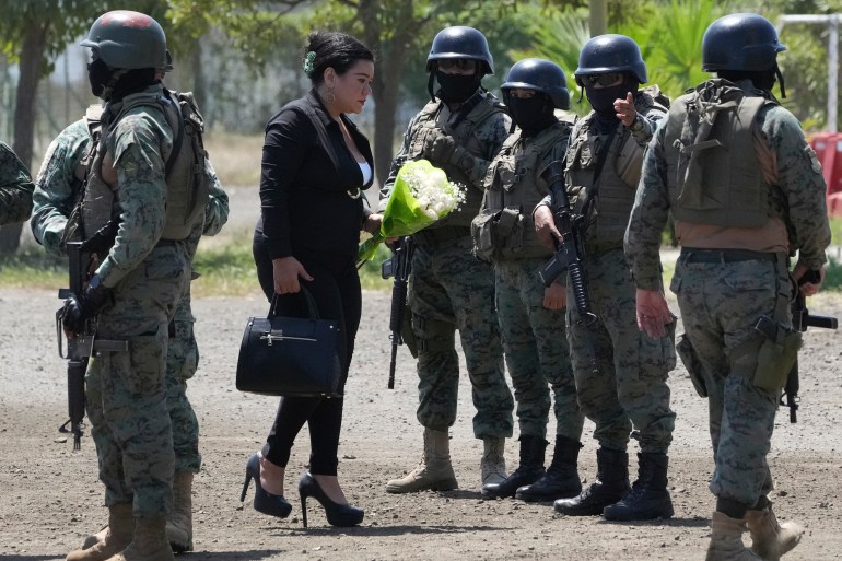 A woman, wearing black, brings a bouquet of white flowers. A line of armed guards in helmets stands in front of her.