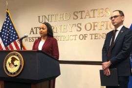 A woman stands at a podium emblazoned with a US seal. Behind her is an American flag and golden words on the wall: "United States Attorney's Office: Western District of Tennessee." A man stands beside her, waiting for his chance at the podium.