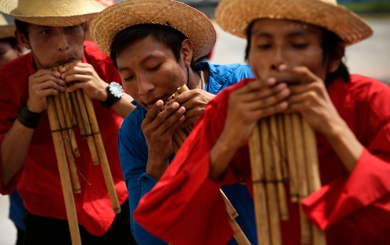 Three teenagers dressed in red with straw hats blow into flutes made of reeds.