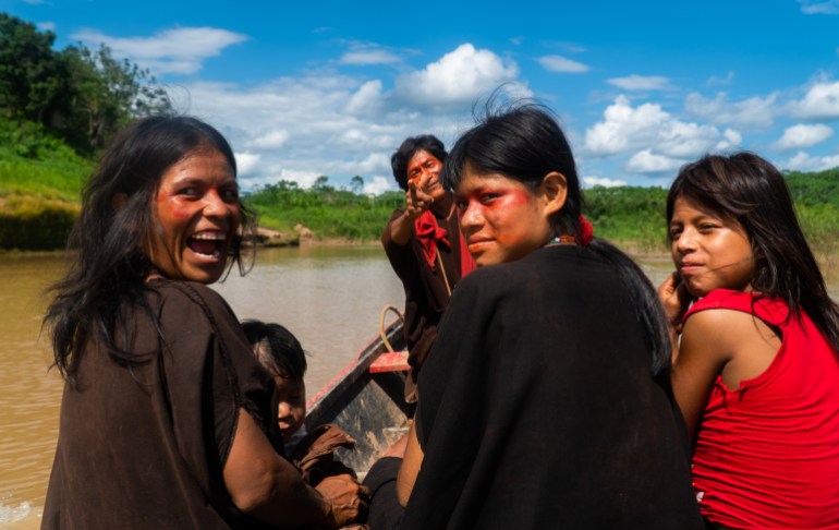 Four women sit in a canoe plying the brown waters of an Amazon river, smiling and looking over their shoulders at the camera.