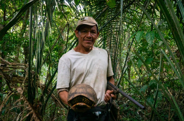 A man in a baseball cap holds out a turtle shell as he stands among palm leaves and bushes.