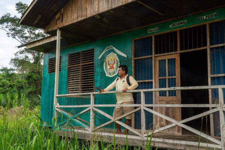 A man stands on the porch of a wooden building, with the seal of Peru painted on the side. The Amazon rainforest is visible in the background, and the building is surrounded by tall grass and shrubs.