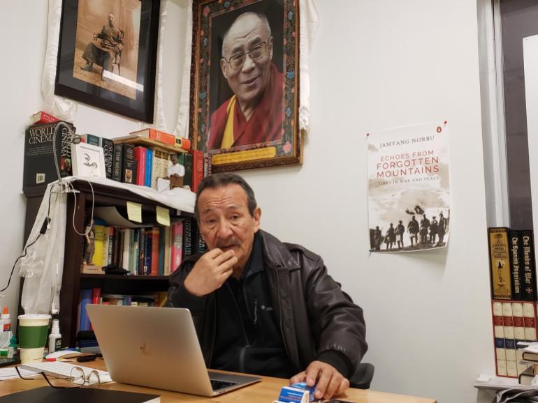 Jamyang Norbu en su escritorio, tiene una computadora abierta frente a él.  Hay un estante de libros a su derecha y un gran retrato del Dalai Lama en la pared detrás de él.  Una maqueta de la portada de su nuevo libro también está en la pared.