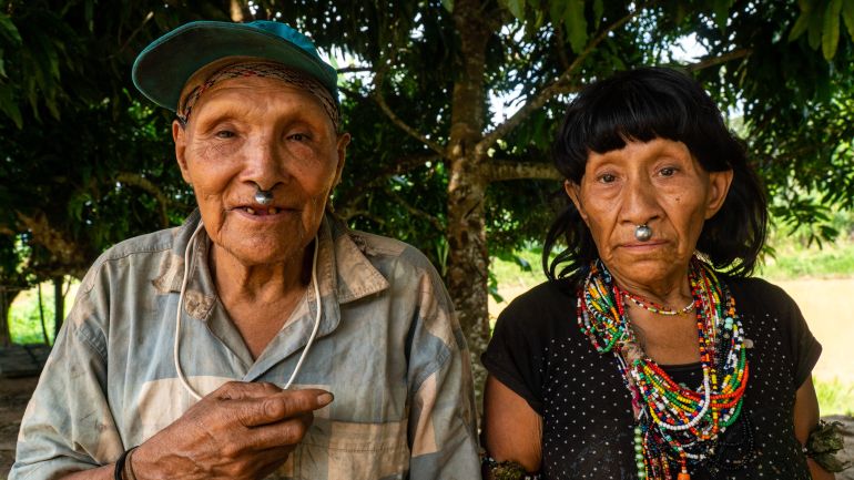 Two Indigenous elders — a man and a woman — sit side by side, looking directly at the camera. They both have what appears to be a metal stud on their upper lip. The woman wears an ornate beaded necklace over her black T-shirt. The man wears a faded blue-and-white striped collared shirt.
