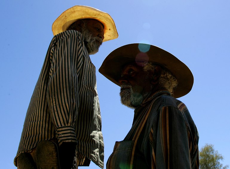 Two Arrernte men silhouetted against a bright blue sky. They are walking past each other