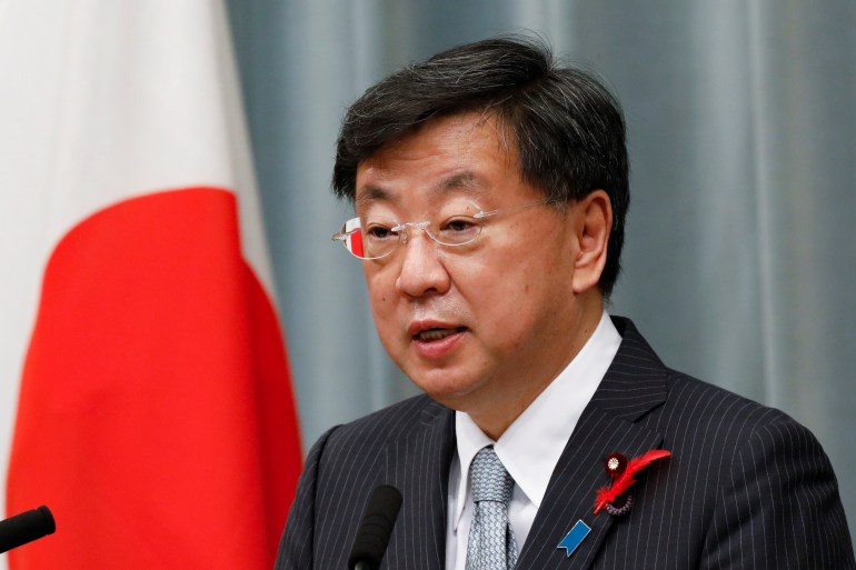 Matsuno Hirokazu, dressed in a suit and light grey tie, speaks at a press conference in front of a Japan flag.