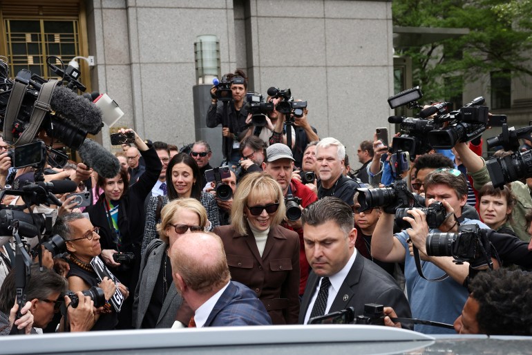 E Jean Carroll — a woman in sunglasses and a bob haircut — plies through a crowd of reporters as she exits the Manhattan Federal Courthouse in May to enter a waiting vehicle.