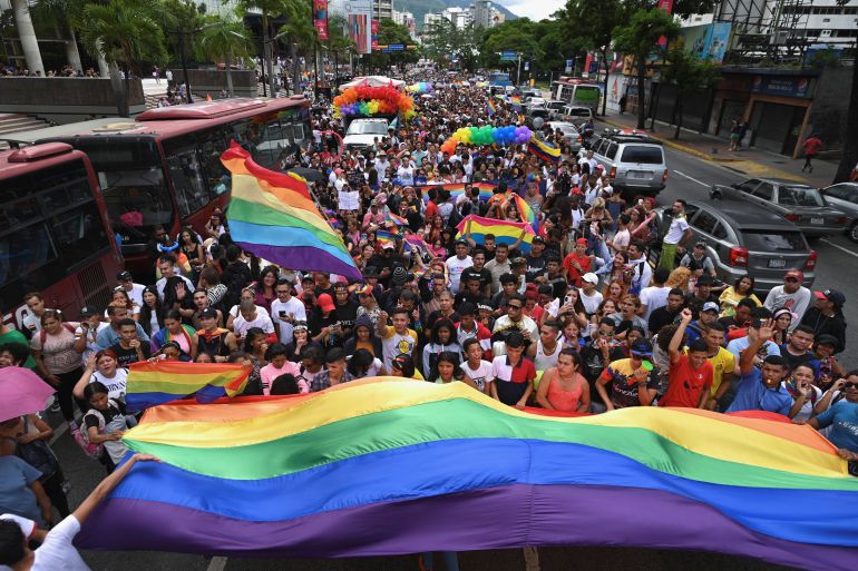A street is crowded by a parade participants waving giant rainbow flags.