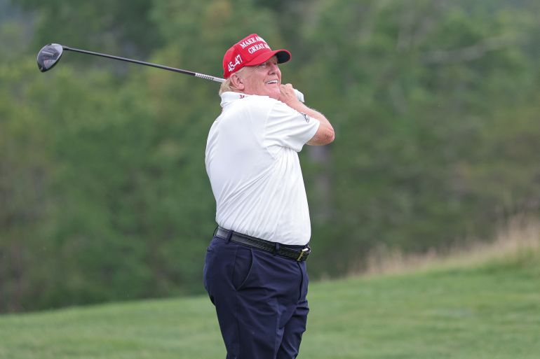 Former President Donald Trump plays his shot from the ninth tee during the ProAm round of the LIV Golf Bedminster golf tournament at Trump National Bedminster. He holds the golf club over his shoulder, having just swung, and he wears a white polo shirt, dark pants and his trademark red baseball cap.