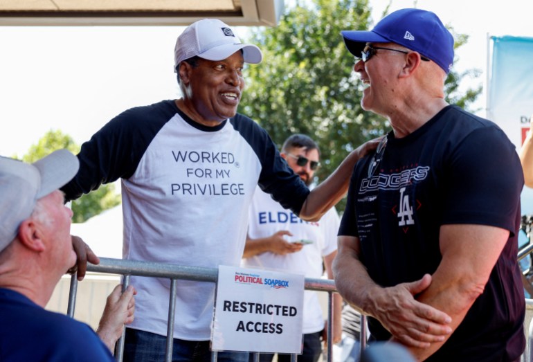 Larry Elder, wearing a baseball cap and a shirt that reads "Worked for my Privilege," pats a man on the shoulder.