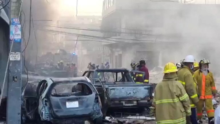 Firefighters and other emergency personnel wearing heavy fire-resistant coats and helmets approach a burning building, surrounded by burnt-out cars.