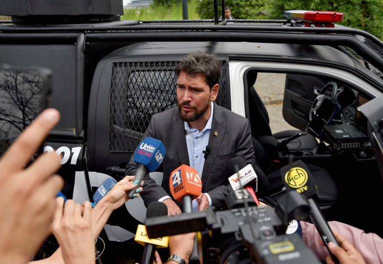 A man, surrounded by microphones, speaks to the press next to a black vehicle with tinted windows.