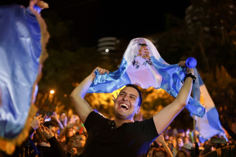Een demonstrant – breed glimlachend – zwaait met een Guatemalteekse vlag boven zijn hoofd terwijl hij feest viert op een druk stadsplein.