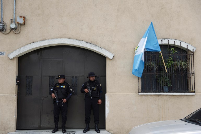 Delante de unas puertas dobles se encuentran dos agentes de policía vestidos de negro y armados.  Una ventana junto a ellos tiene una bandera guatemalteca ondeando entre las rejas de seguridad.