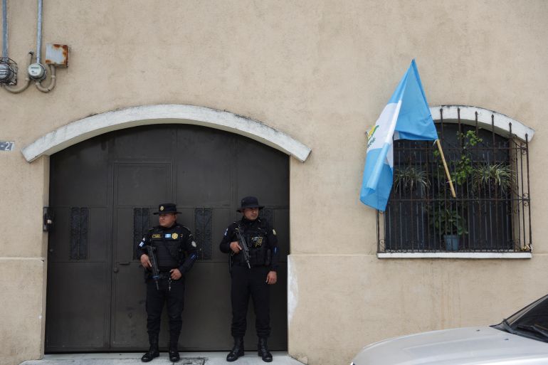 Outside a set of double doors, two police officers dressed in black and carrying guns stand. A window next to them has a Guatemalan flag waving from between its security bars.