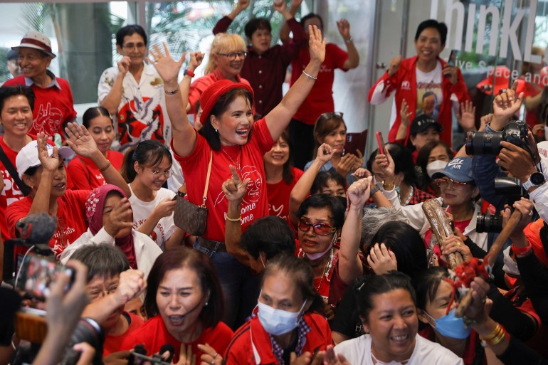 Pheu Thai supporters wearing red shirts celebrate Srettha's victory. One woman has her arms outstretched, others are punching the air. They look delighted.