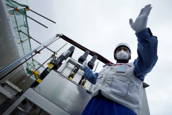 A company official in a hazmat suit at the Fukushima plant. He has his arm held out and is explaining something. There are pipes and tanks behind him