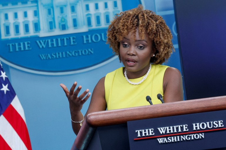 Karine Jean-Pierre speaks from the White House podium, wearing a sleeveless lime green shift dress. Next to her is a US flag, and behind her is the seal of the White House. 