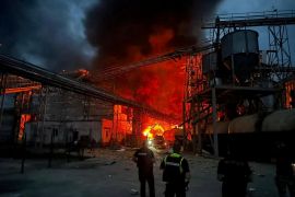 A vegetable oil factory on fire after a Russian missile attack. There are flames and black smoke. There are two rescue workers at the scene.