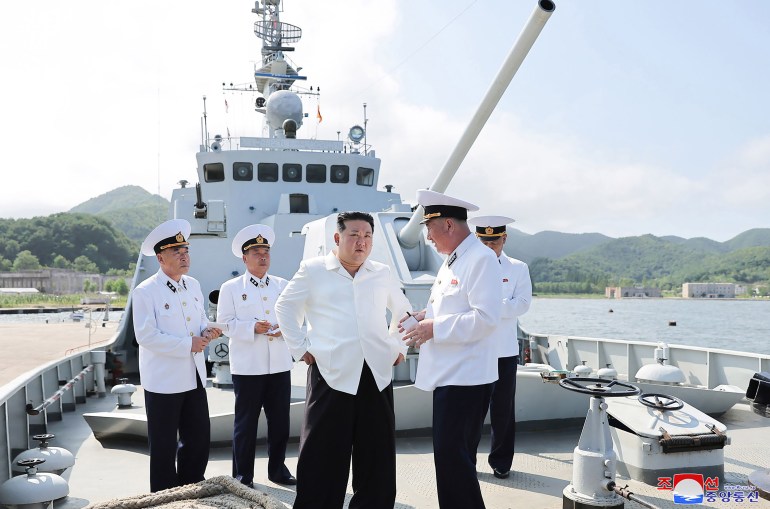 Kim Jong Un no convés de um navio naval.  Oficiais da marinha estão atrás dele.  Eles estão vestindo camisas brancas e calças pretas.