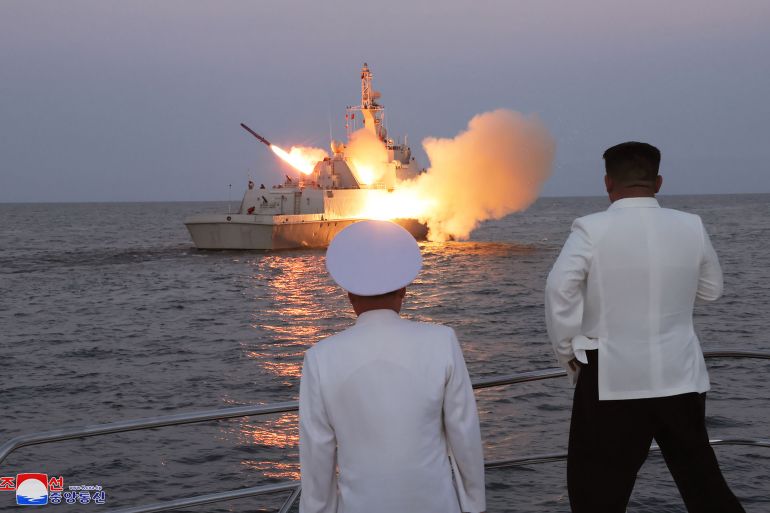 Alevler içindeki bir tekneden bir füze fırlatıldı.  Kim Jong Un ve bir donanma subayı iskeleden izliyor.  Sırtları kameraya dönük.