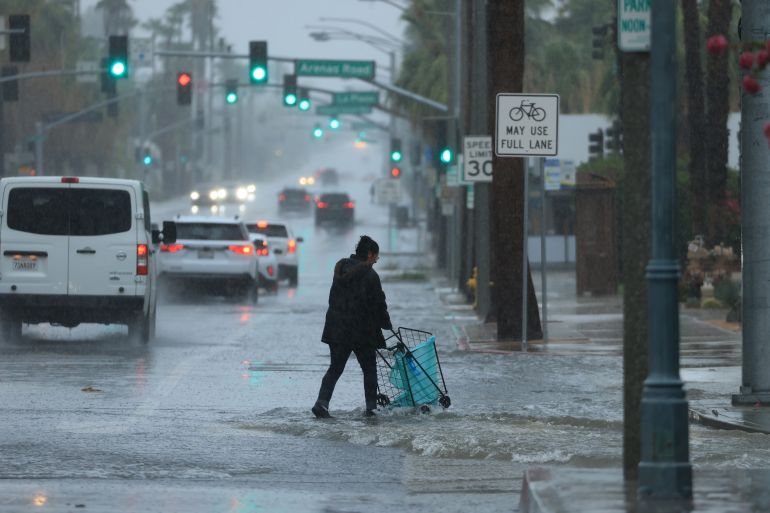 A pedestrian walks across a water covered street with a trolley. Cars are queued up along the road. The traffic lights are illuminated in the low light. There is lots of rain.