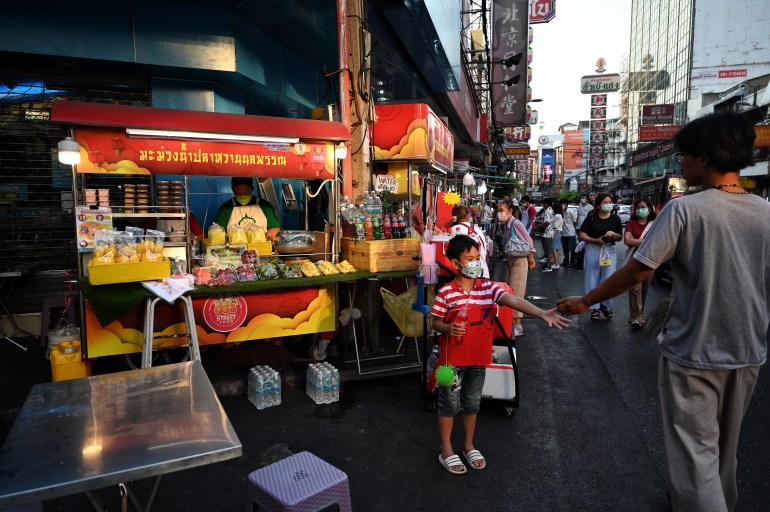 A photo of street food stalls in Bangkok, Thailand with a food stall selling mangoes and people walking in the street.