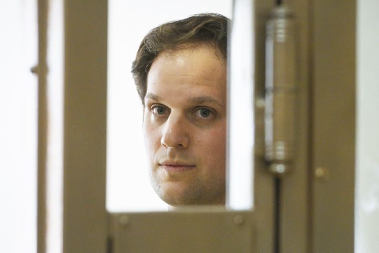 A man, Evan Gershkovich, peers out from a glass cage as he attends a hearing in June.