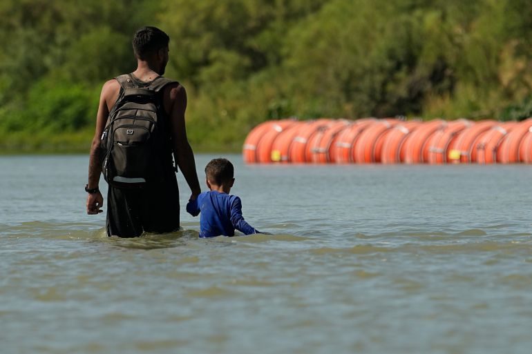 A migrant standing in the river stares at a floating border barrier
