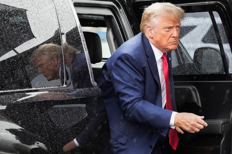 Donald Trump, dressed in a blue suit and red tie, exits a black vehicle with tinted windows.