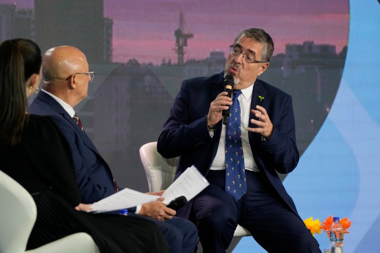 A man in a dark suit and glasses, Bernardo Arevalo, sits on a white chair and speaks into a microphone, his arms lifted slightly in gesture. Interviewers sit across from him on the stage.