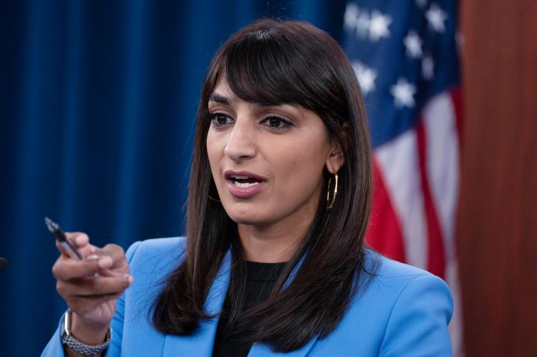 A woman in a blue suit speaks and points with a pen. Behind her is a United States flag.