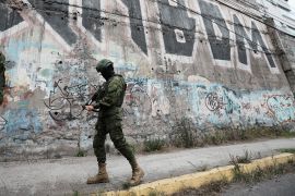 A soldier in fatigues, a helmet and a flak jacket walks with a gun down a sidewalk lined with graffiti.