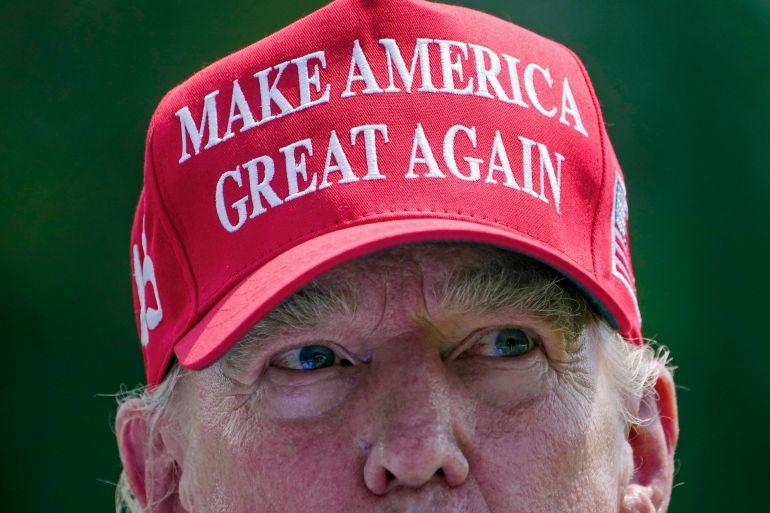 Um close da metade superior da cabeça de Donald Trump.  Ele usa seu boné vermelho, sua marca registrada, que diz: "Torne a América grande novamente."