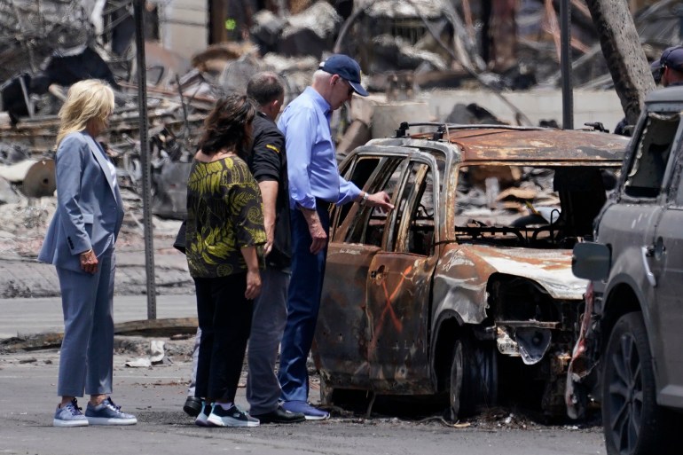 Joe Biden reaches out to touch a burned car, its windows gone and its exterior a mottle of grey and rust colors. Behind him are members of the community and his wife, Jill Biden.