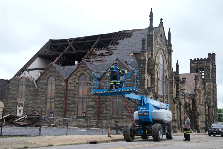 A Gothic-style church is seen with massive tears in its roof. A blue crane with workers in its basket is seen parked in the front.