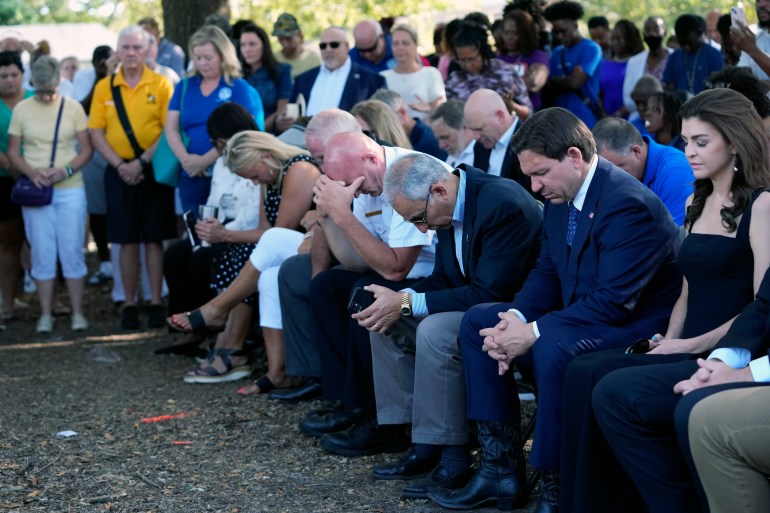 Florida Governor Ron DeSantis (seated second from right) prays during the vigil for the Jacksonville attack. He is seated outside with other officials. HIs wife is next to him. Their heads are bowed.