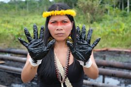An Indigenous woman, her head crowned in yellow feathers, raises two hands covered in black tar