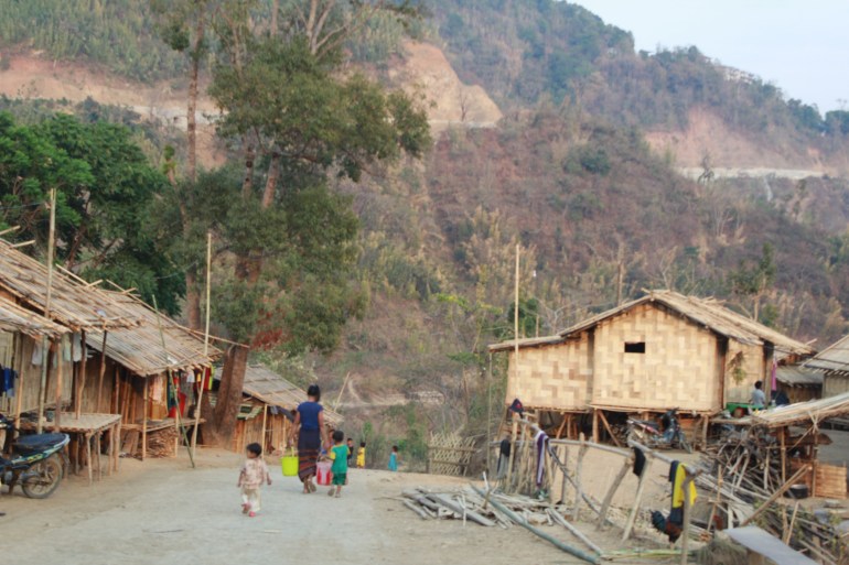 A view along a street at a displacement camp for Myanmar people in Mizoram. The buildings are woven from bamboo and palm. It looks quite dry and dusty, a woman is walking away with two children.