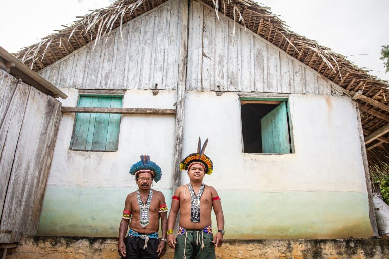 Two Indigenous men — a father and son — stand in front of a house with white walls and a palm-thatched roof.