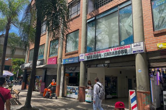 The exterior of a building in Medellin Colombia, that has a sign that reads: "Centro de Negocios y Servicios. San Juan-Carabobo." Palm trees are planted on the sidewalk outside.